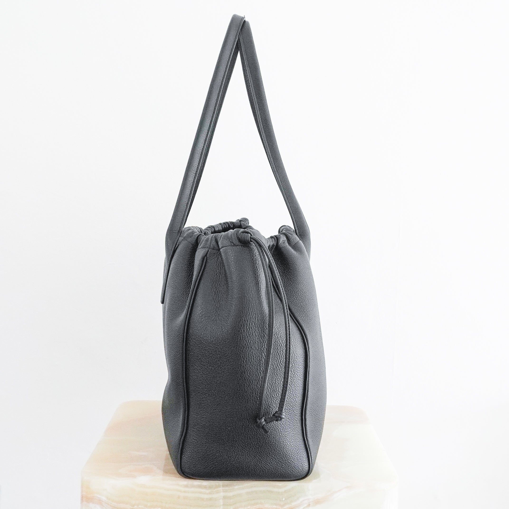 Side photo Celine Cuir Triomphe Cabas Drawstring Tote, crafted from luxurious black grained calfskin leather and featuring the house's iconic oversized embossed Triomphe logo to the front.