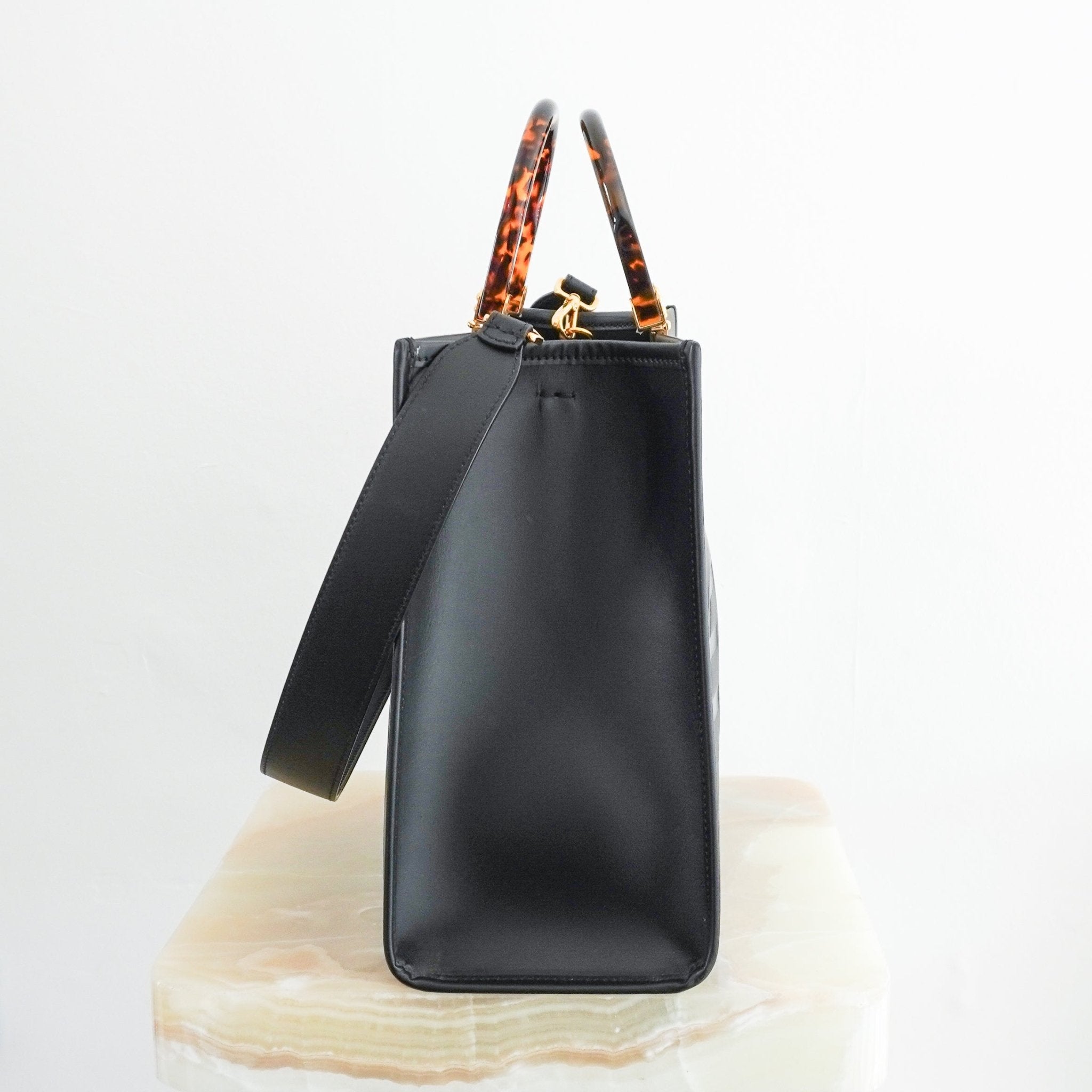 Side view of a structured black leather tote bag with tortoiseshell-effect top handles, gold-tone hardware, and a detachable shoulder strap.