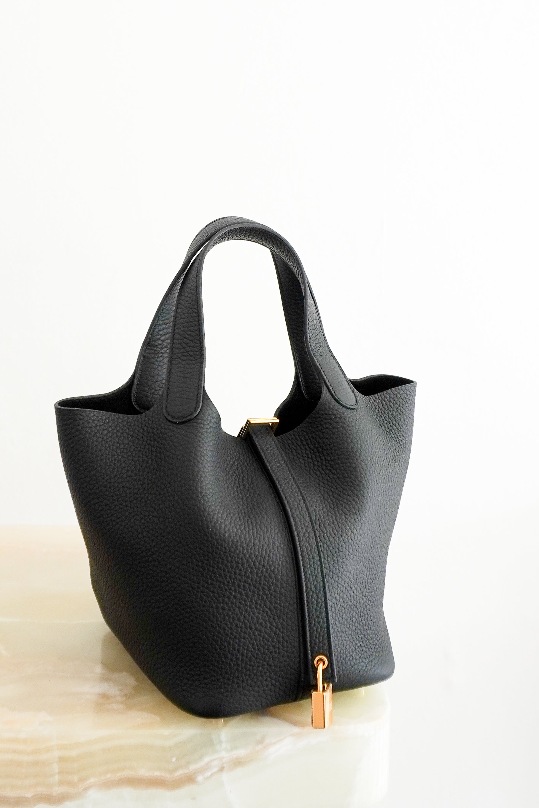 Front picture of a Hermès Picotin Lock 22 tote bag in black pebbled Clemence leather with gold-tone hardware and a signature padlock closure, photographed on a cream marble pedestal.