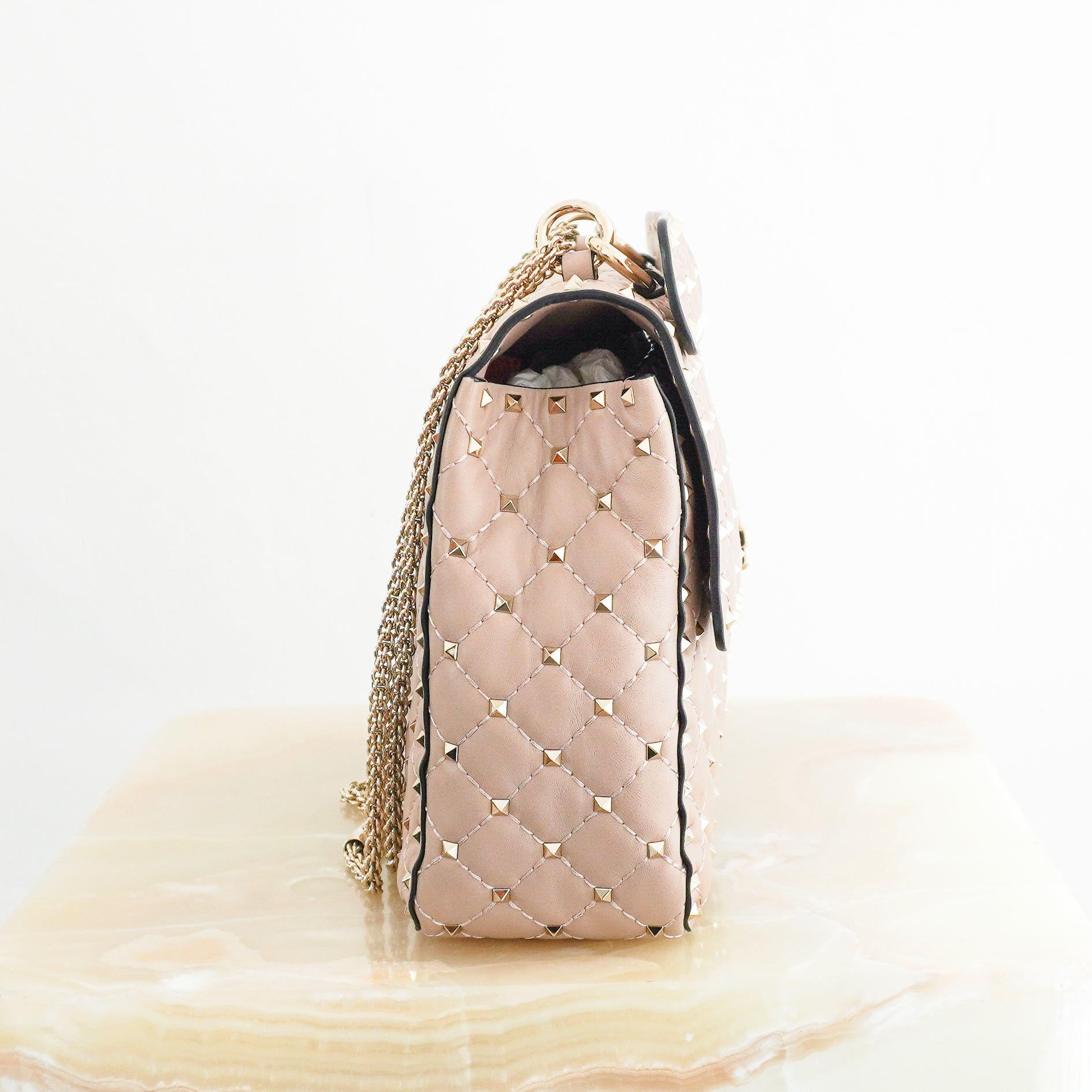 Side view of a Valentino Garavani blush pink quilted leather shoulder bag with gold-tone stud detailing and a structured flap closure.