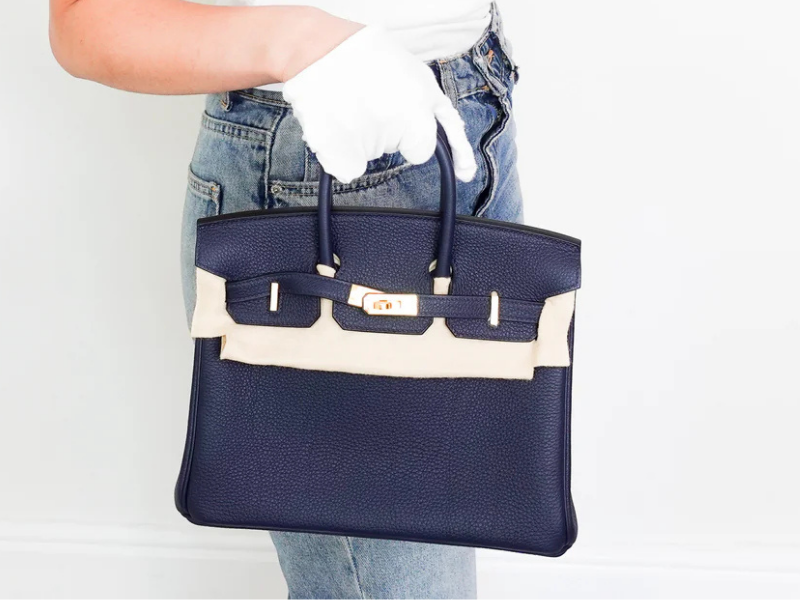 A woman holding two luxury Hermès handbags—a light blue Hermès Kelly with a decorative silk Twilly wrapped around the handle and a deep teal grained leather Hermès Birkin with gold hardware—at the Reloved Again Chelsea boutique.