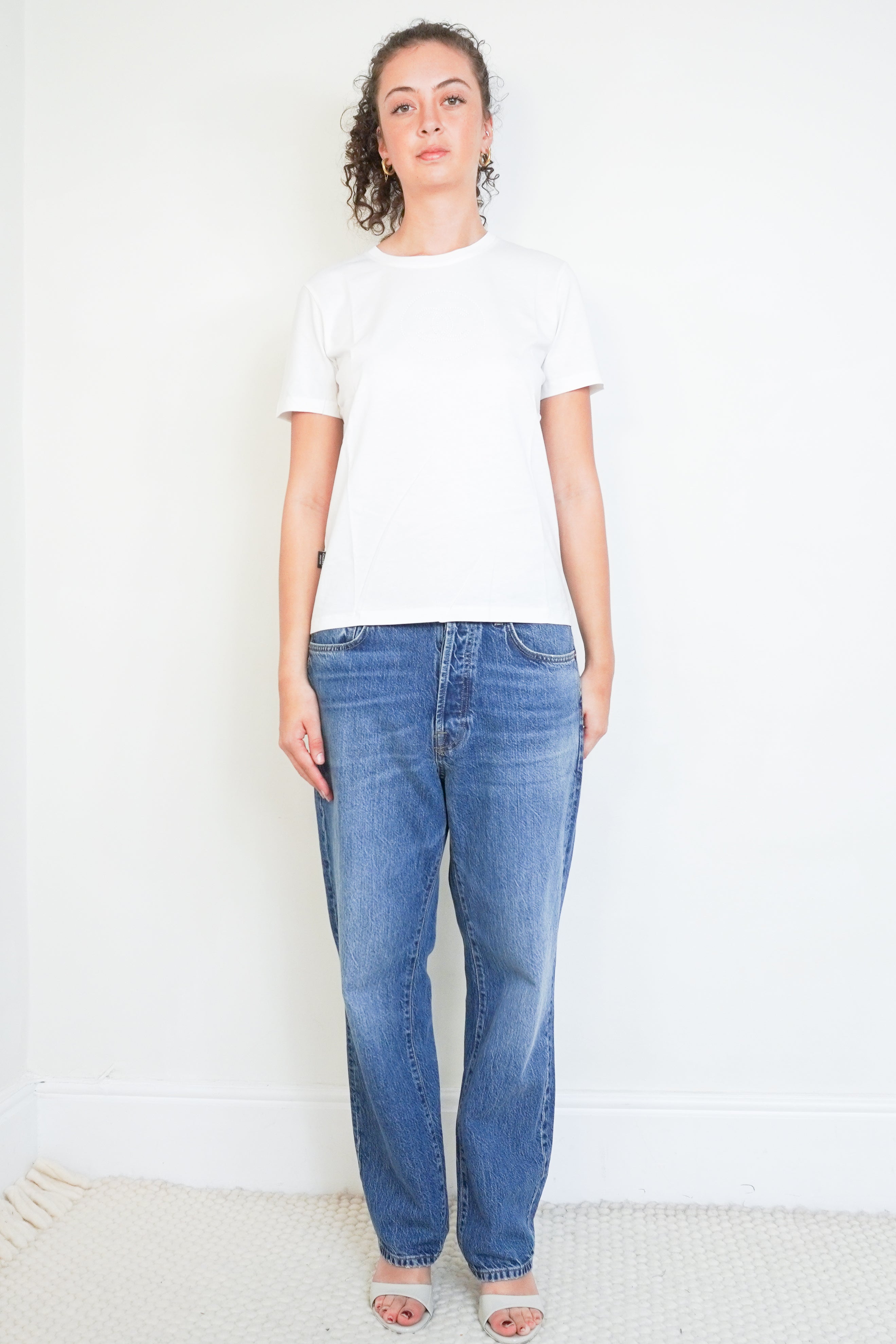 Front photo of a Chanel white short-sleeved T-shirt featuring a subtle CC crystal logo detail on the side hem, paired with blue jeans.