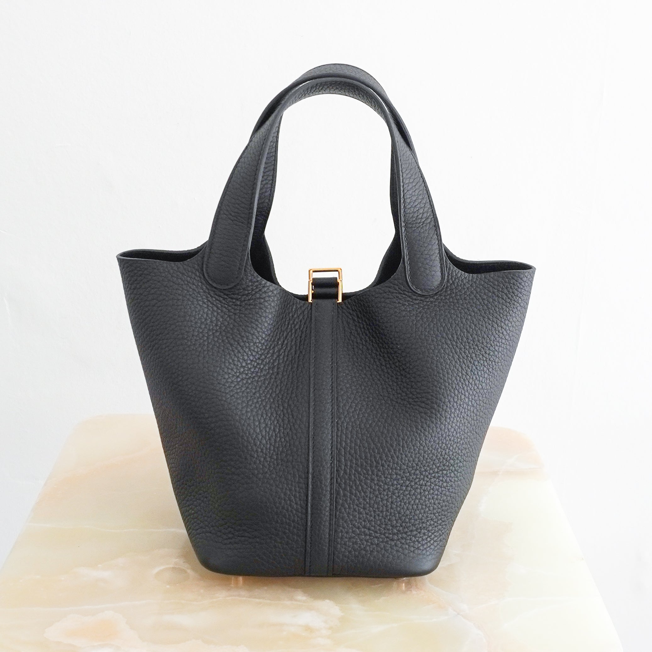 Back picture of a Hermès Picotin Lock 22 tote bag in black pebbled Clemence leather with gold-tone hardware and a signature padlock closure, photographed on a cream marble pedestal.