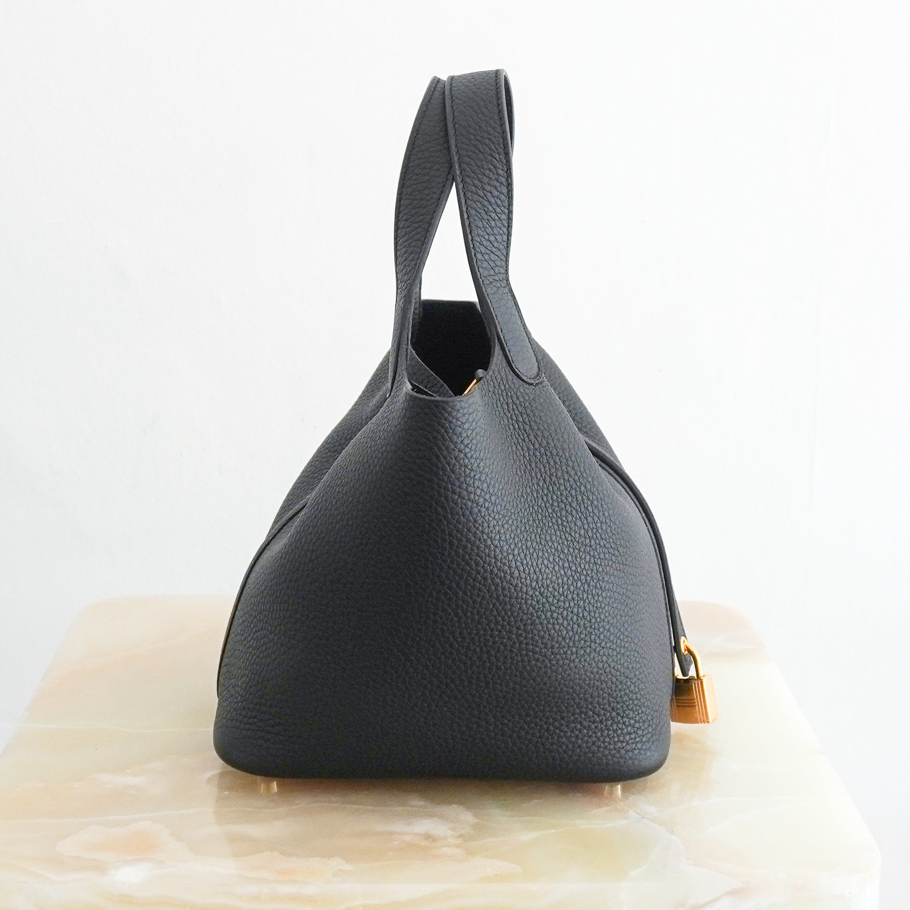 Side picture of a Hermès Picotin Lock 22 tote bag in black pebbled Clemence leather with gold-tone hardware and a signature padlock closure, photographed on a cream marble pedestal.
