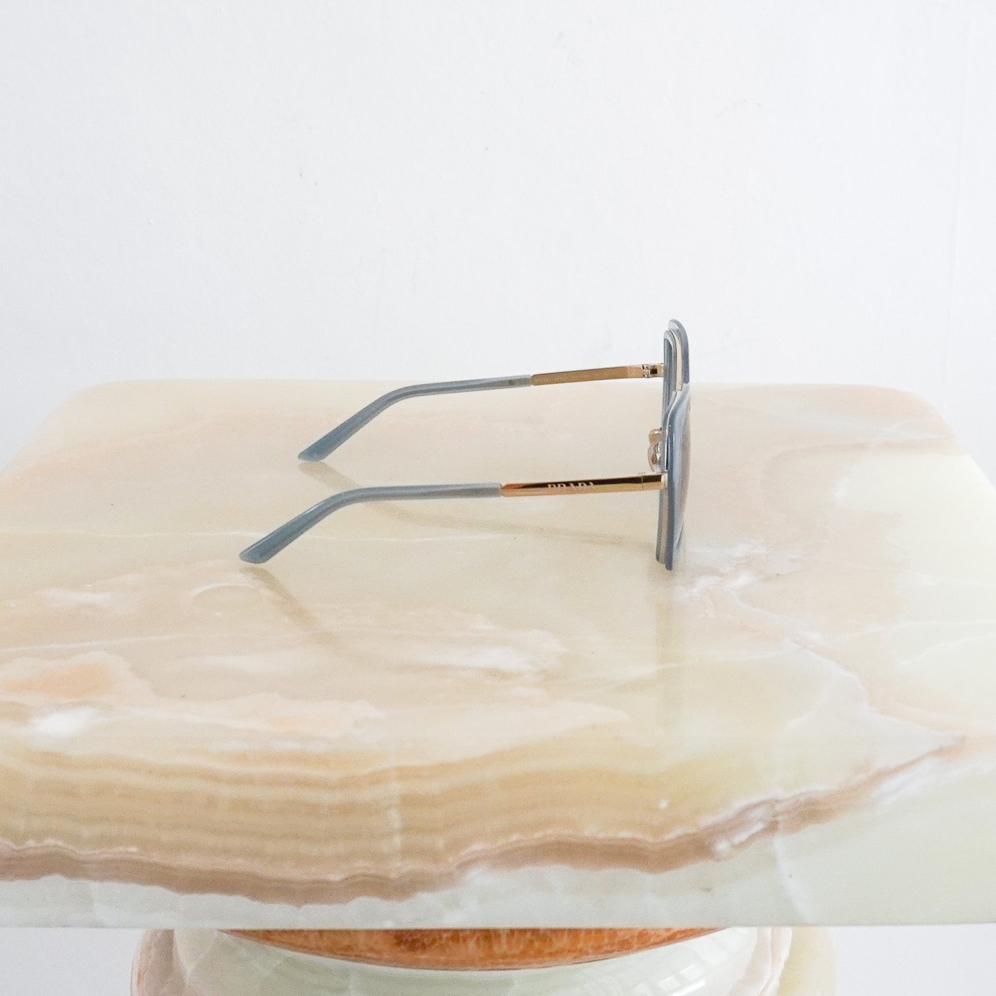 Side profile of Prada sunglasses featuring light blue frames and gold-tone hardware arm details.