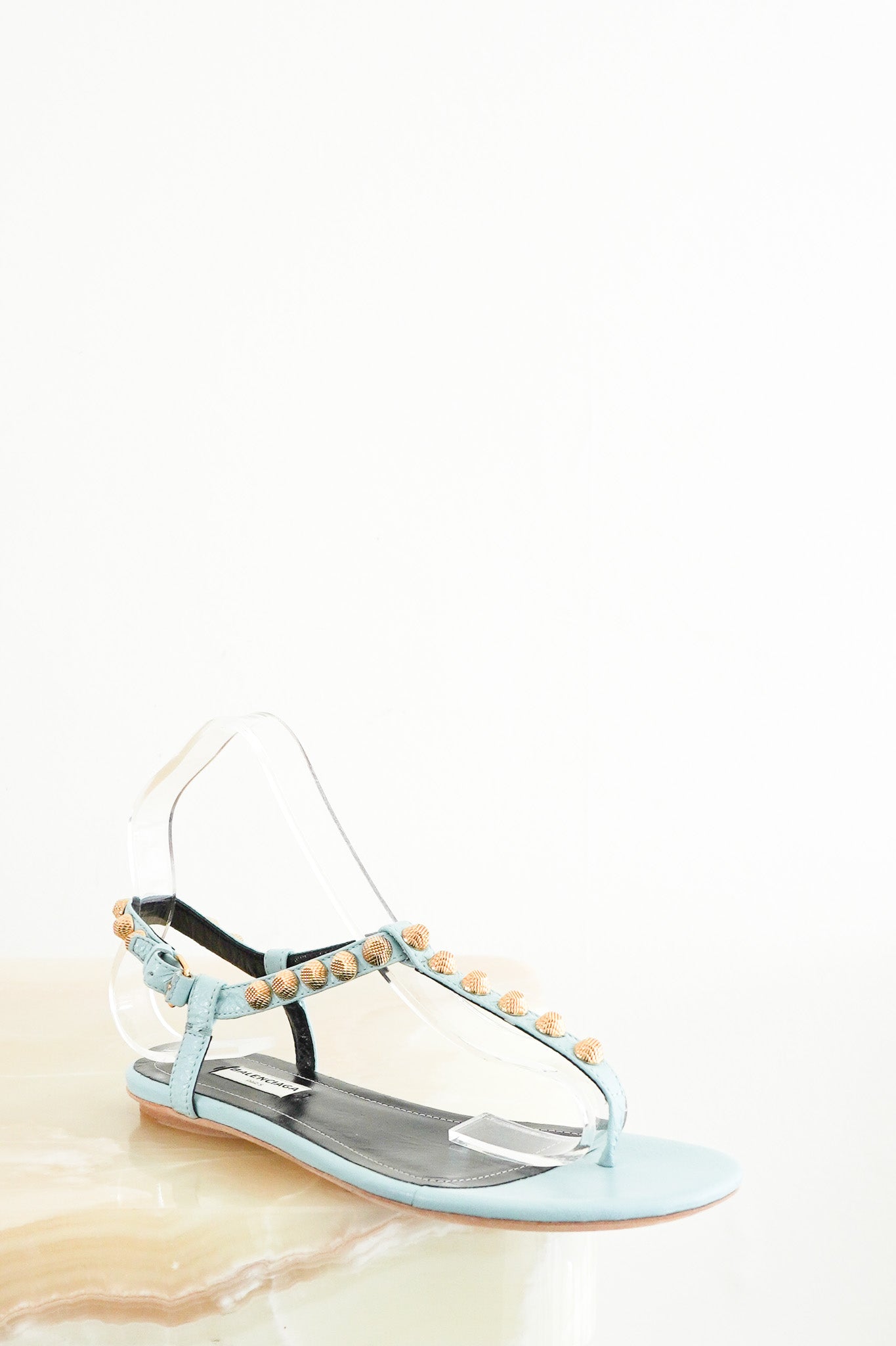 Front photo of Balenciaga Blue Arena Sandals, featuring light blue leather t-strap bands adorned with textured gold-tone studs, a flat sole, and an adjustable ankle strap.
