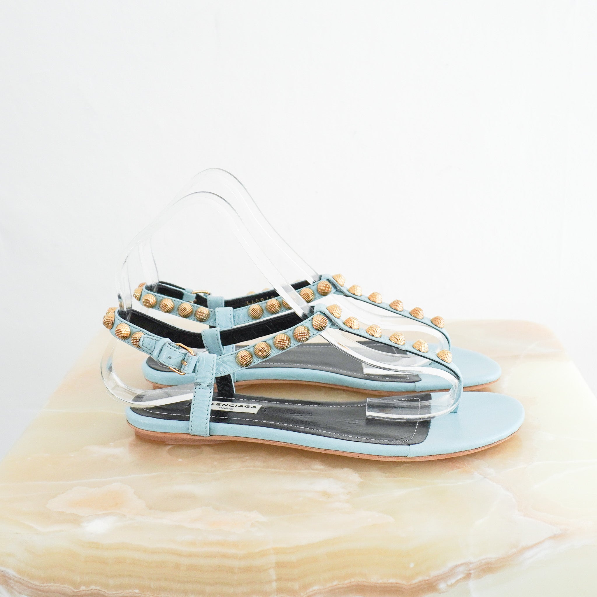 Side photo of Balenciaga Blue Arena Sandals, featuring light blue leather t-strap bands adorned with textured gold-tone studs, a flat sole, and an adjustable ankle strap.